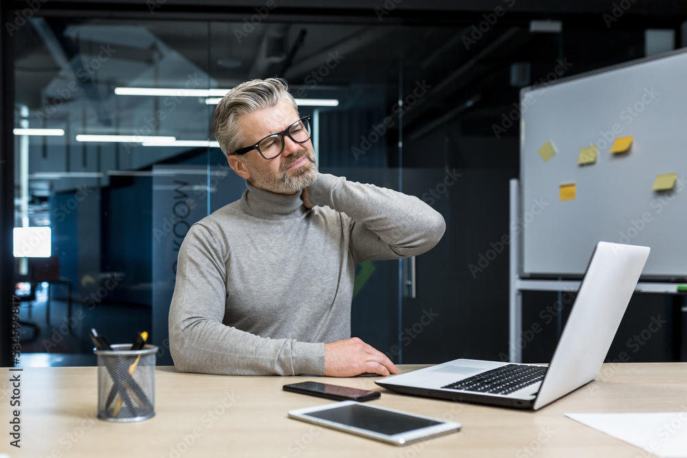A tired and exhausted businessman man in glasses sits in the office at the desk, holds his neck, massages his neck with his hand. He feels pain in his muscles. A tired and exhausted businessman man in glasses sits in the office at the desk, holds his neck, massages his neck with his hand. He feels pain in his muscles.