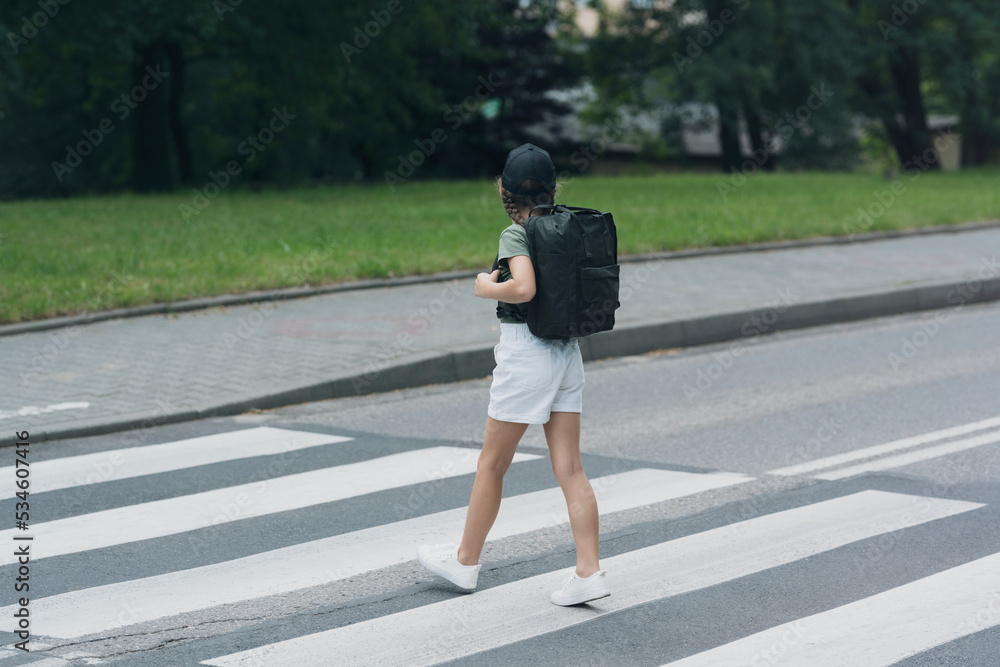 Fototapeta premium A girl with braids in a black backpack hat crosses the streets at the pedestrian crossing.
