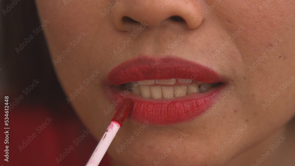 Naklejka premium CLOSE UP: Detailed shot of young woman applying red lip gloss to her lips
