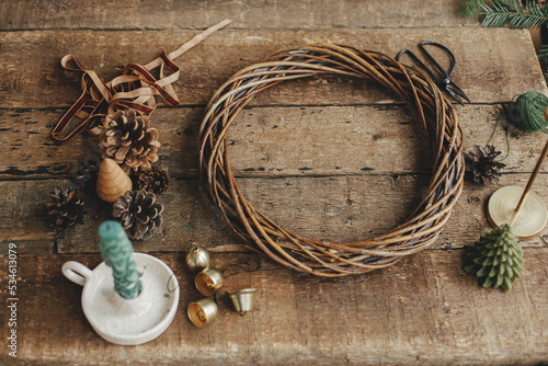 Making rustic Christmas wreath. Round wicker wreath, pine cones, ribbon, golden bells and candles on rustic wooden table. Christmas still life, holiday preparations