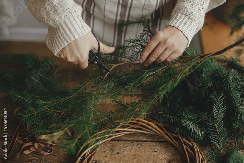 Making rustic Christmas wreath. Female hands cutting fir branches with scissors on background of rustic wooden table with ribbon. Moody Christmas image, holiday preparations