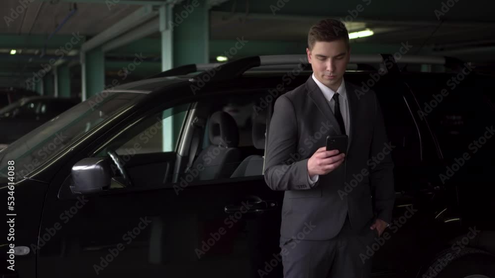 A young businessman uses a phone in a car park. A man in a suit holds a phone in his hand. A young businessman uses a phone in a car park. A man in a suit holds a phone in his hand.