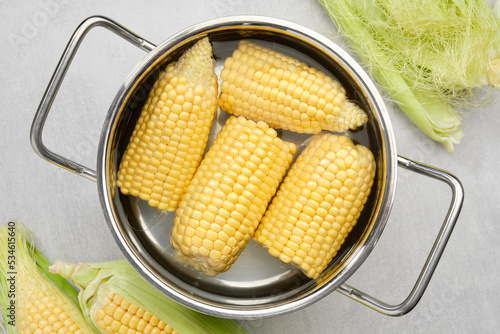 Cooking pot of yellow sweet corn cobs prepared for boiling, raw corncobs for cooking on kitchen table, top view.