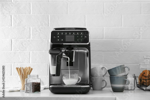 Modern coffee machine pouring milk into glass cup on white countertop in kitchen