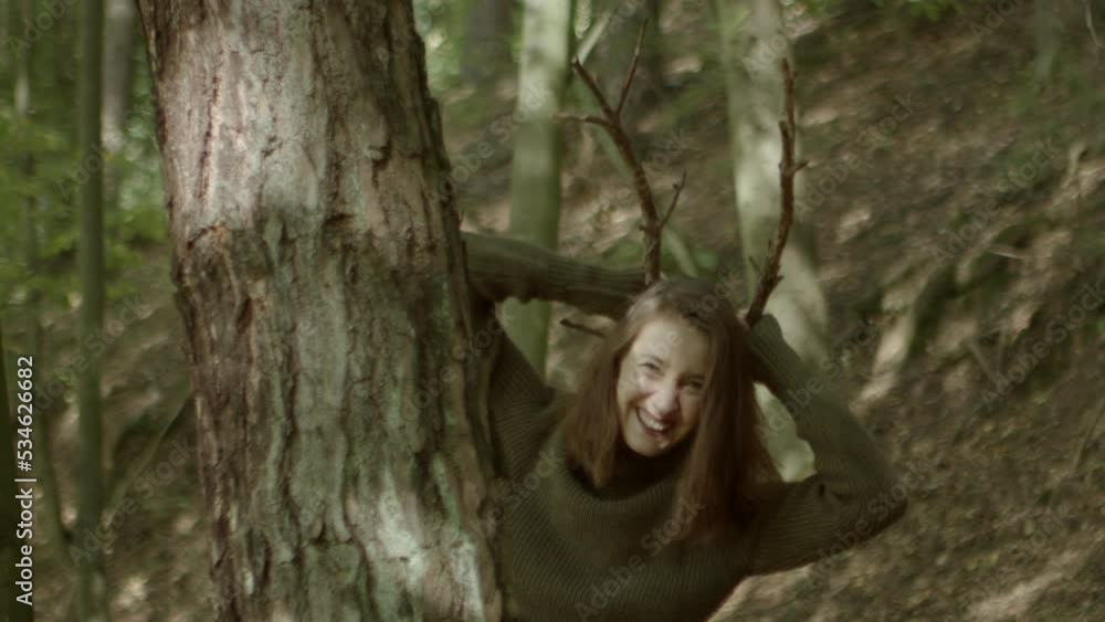 Young woman peeks out from behind a tree, puts two sticks on her head and pretends to be a deer. Young woman peeks out from behind a tree, puts two sticks on her head and pretends to be a deer.