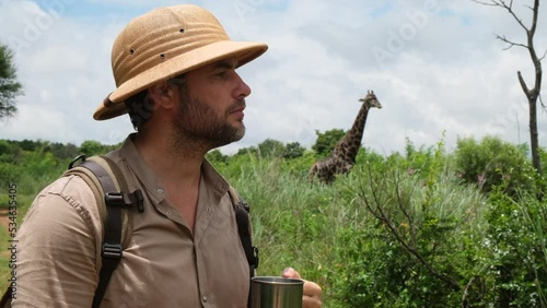 a man in a safari hat drinks water near a giraffe. Happy man traveller on a safari in Africa, travels by car in Kenya and Tanzania, watches life wild giraffes, zebras and antelopes in the savannah. photo