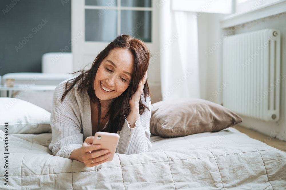 Young woman forty year with brunette long hair in cozy knitted cardigan using mobile phone in bed at home Young woman forty year with brunette long hair in cozy knitted cardigan using mobile phone in bed at home