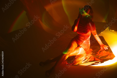 Glamorous portrait of a young beautiful blonde in a stylish scarlet short dress, sitting on the floor, refraction effects of colored light, futuristic fashion shooting in neon light