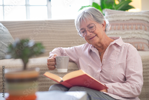 Smiling attractive senior woman relaxing sitting on the floor reading a book holding a cup of coffee or tea. Caucasian mature lady studying, learning at home