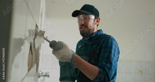 A man in work clothes wearing a shirt and cap is hammering tiles in the kitchen bathroom doing renovations in the house.