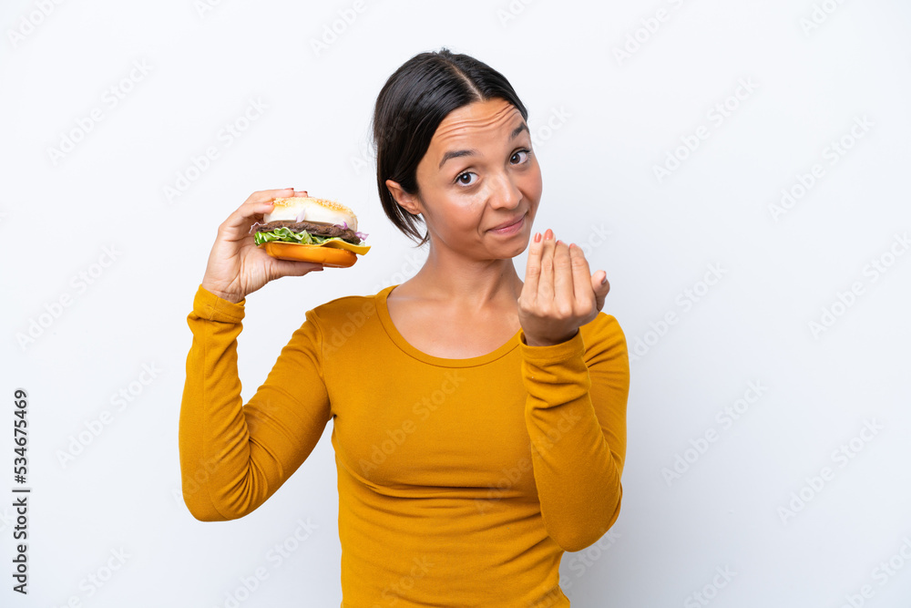 Young hispanic woman holding a burger isolated on white background inviting to come with hand. Happy that you came Young hispanic woman holding a burger isolated on white background inviting to come with hand. Happy that you came