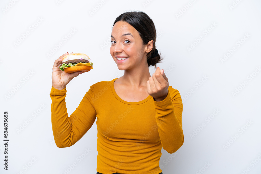 Young hispanic woman holding a burger isolated on white background making money gesture Young hispanic woman holding a burger isolated on white background making money gesture