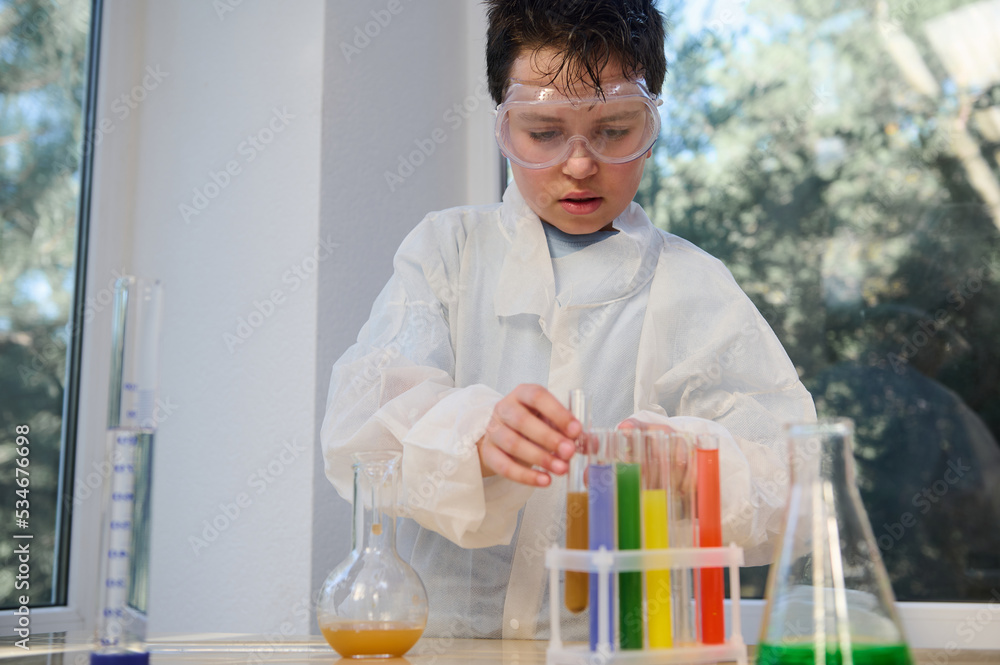 Confident smart Caucasian boy wearing safety goggles and white lab coat