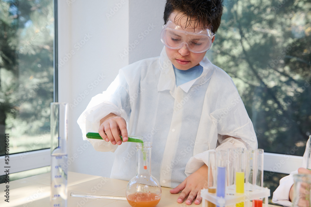 Smart schoolboy in protective lab gear, standing at table in the school science laboratory, pouring chemical liquid from a test tube into a glass flask, conducting experiments while learning Chemistry Smart schoolboy in protective lab gear, standing at table in the school science laboratory, pouring chemical liquid from a test tube into a glass flask, conducting experiments while learning Chemistry