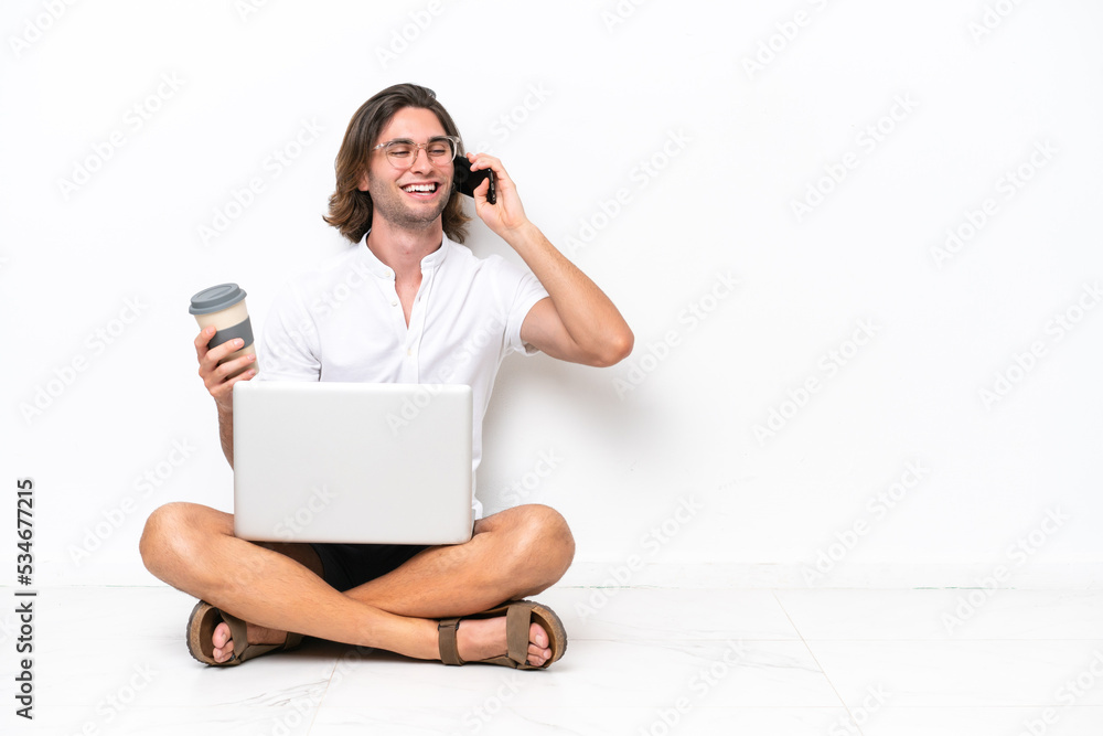 Young handsome man with a laptop sitting on the floor isolated on white background holding coffee to take away and a mobile Young handsome man with a laptop sitting on the floor isolated on white background holding coffee to take away and a mobile