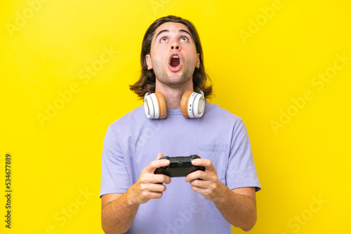 Young handsome caucasian man playing with a video game controller over isolated on yellow background looking up and with surprised expression
