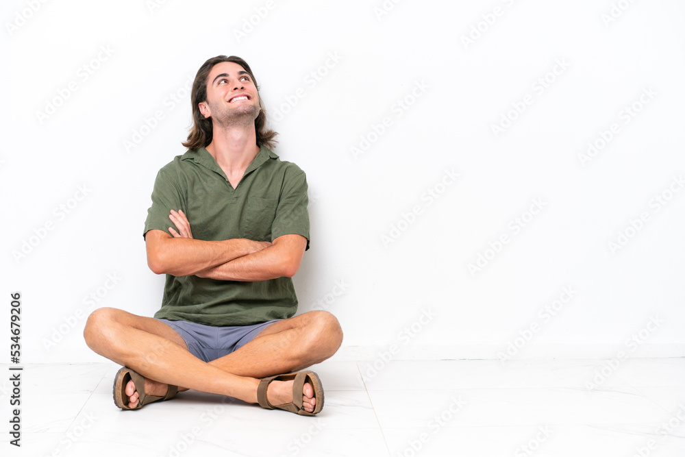 Young handsome man sitting on the floor isolated on white background looking up while smiling Young handsome man sitting on the floor isolated on white background looking up while smiling