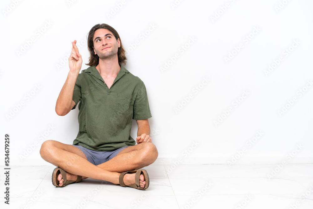 Young handsome man sitting on the floor isolated on white background with fingers crossing and wishing the best Young handsome man sitting on the floor isolated on white background with fingers crossing and wishing the best
