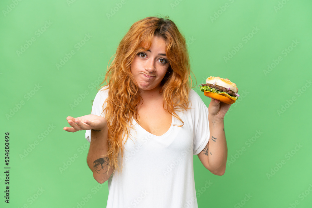 Young caucasian woman holding a burger isolated on green screen chroma key background making doubts gesture while lifting the shoulders Young caucasian woman holding a burger isolated on green screen chroma key background making doubts gesture while lifting the shoulders