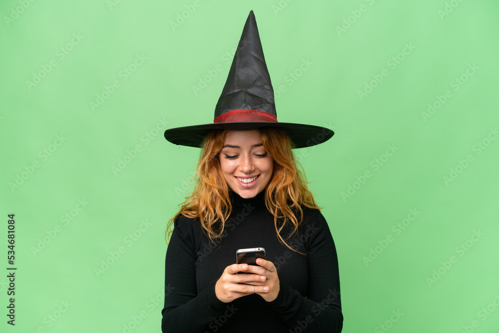 Young caucasian woman costume as witch isolated on green screen chroma key background sending a message with the mobile