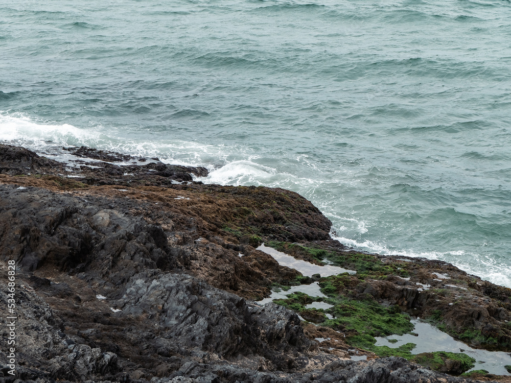 Obraz premium Cold sea water and coastal rocks. Cloudy weather on the Atlantic coast. Surf and white sea foam, rock formation beside body of water.