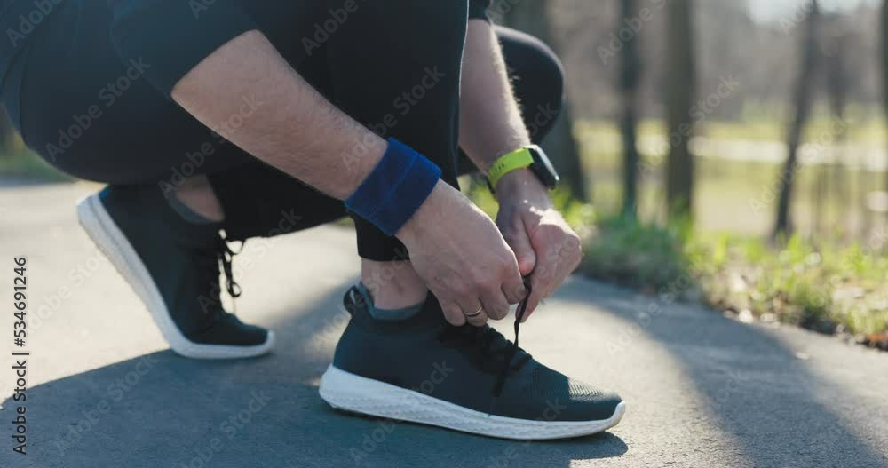 Close-up of man tying shoelaces. He is dressed in black sportswear and has black sneakers with white soles. The man has wristband and watch with green strap on hand. There is wedding ring. Close-up of man tying shoelaces. He is dressed in black sportswear and has black sneakers with white soles. The man has wristband and watch with green strap on hand. There is wedding ring.