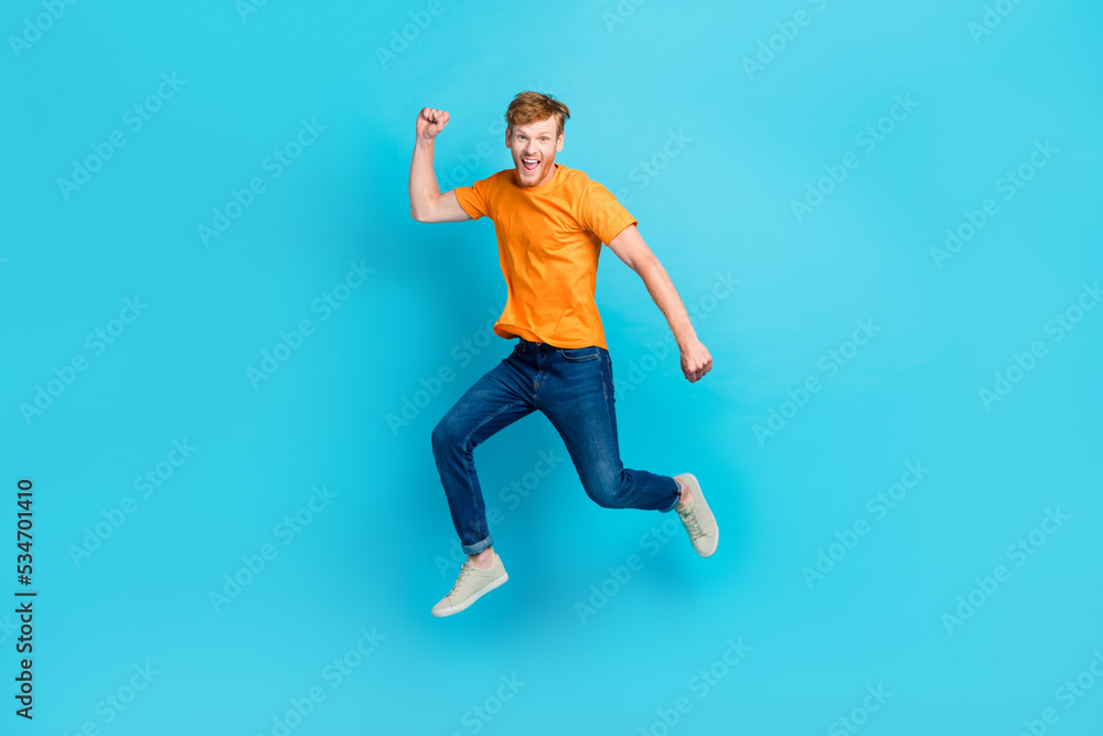 Full length photo of hooray ginger hair guy jump wear t-shirt jeans sneakers isolated on blue color background Full length photo of hooray ginger hair guy jump wear t-shirt jeans sneakers isolated on blue color background