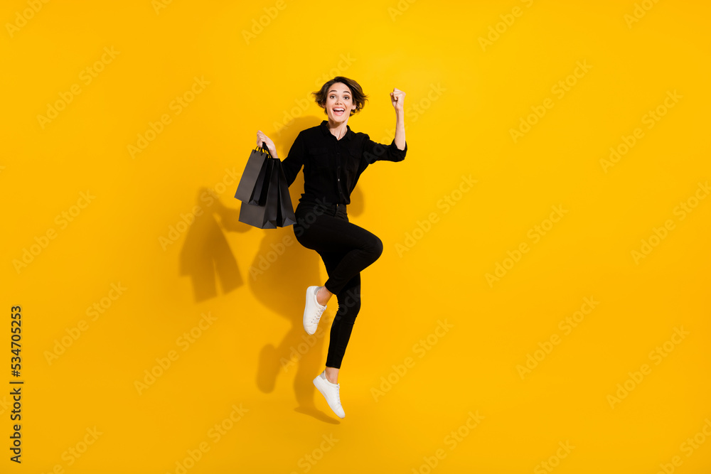 Photo of positive lucky girl dressed formal shirt jumping high rising fist bags empty space isolated yellow color background