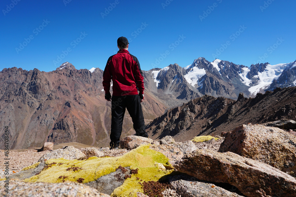 The guy looks at the mountains and snow-capped peaks. High mountains stretch to the blue sky. The peaks of the mountains are covered with glaciers. Steep cliffs down. There are large stones and moss