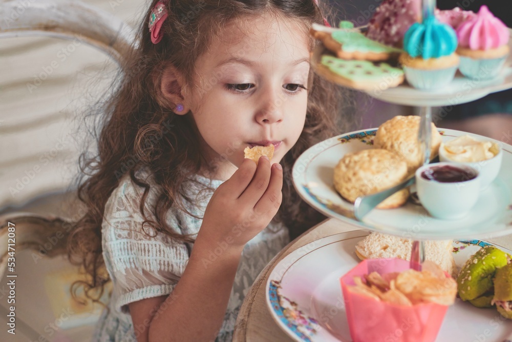 A cute little girl enjoying high tea with ceramic tiered plate stand with scones, cupcakes, sandwiches and crisps