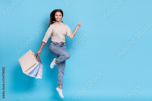 Full size photo of cool young lady jump with bags point promo wear shirt jeans shoes isolated on blue color background