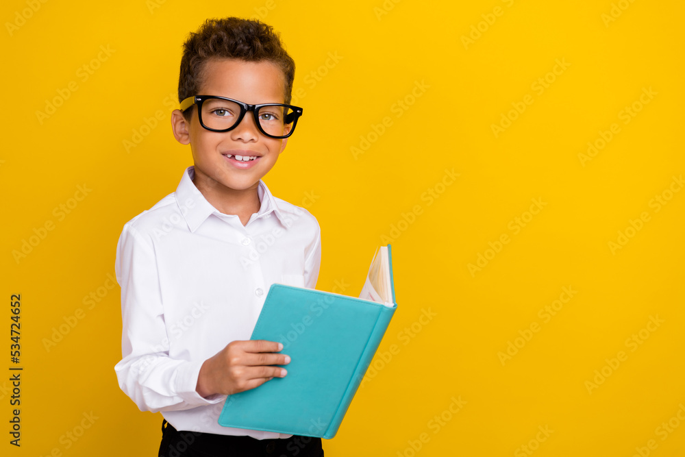 Photo of clever cute boy learner wear eyeglasses hold copybook read task question exercise empty space isolated on yellow color background Photo of clever cute boy learner wear eyeglasses hold copybook read task question exercise empty space isolated on yellow color background