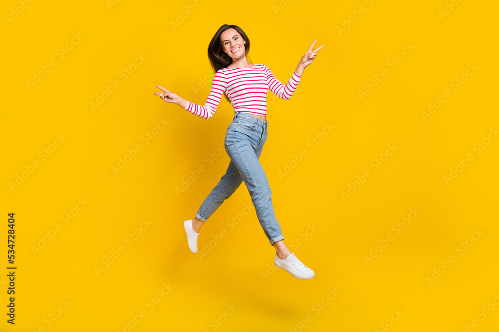 Full size photo of gorgeous good mood girl wear striped long sleeve jeans showing v-sign jump fly isolated on yellow color background