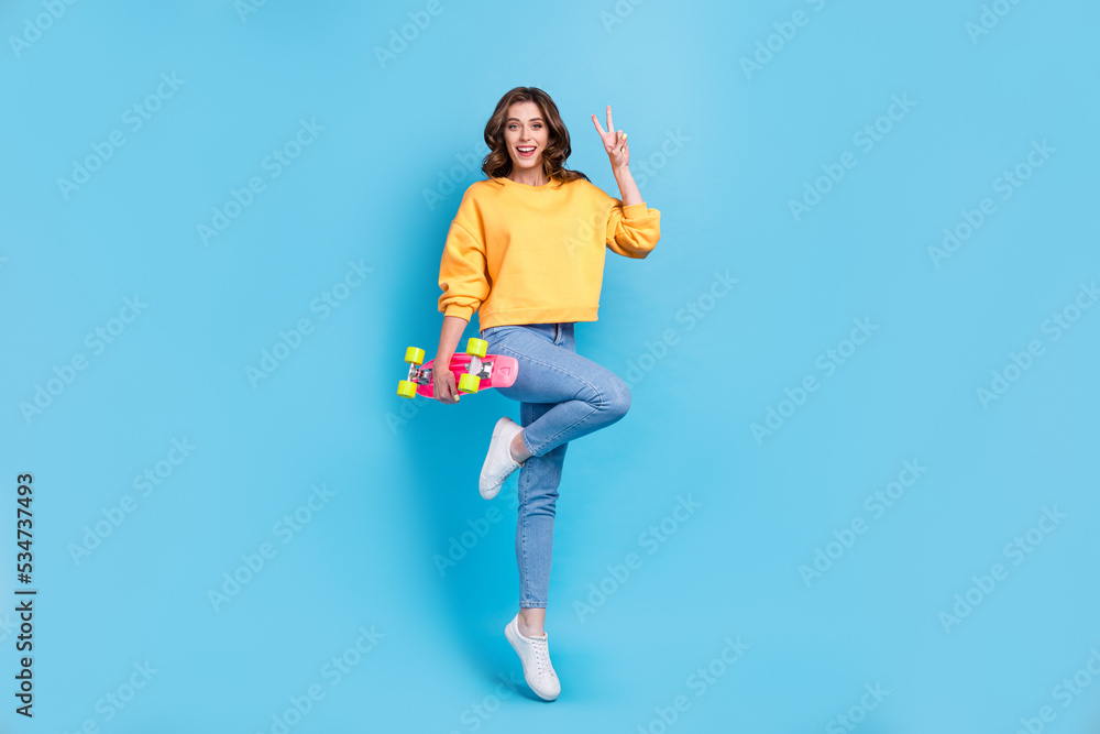 Full length photo of charming funny lady wear yellow sweater jumping high holding longboard showing v-sign isolated blue color background