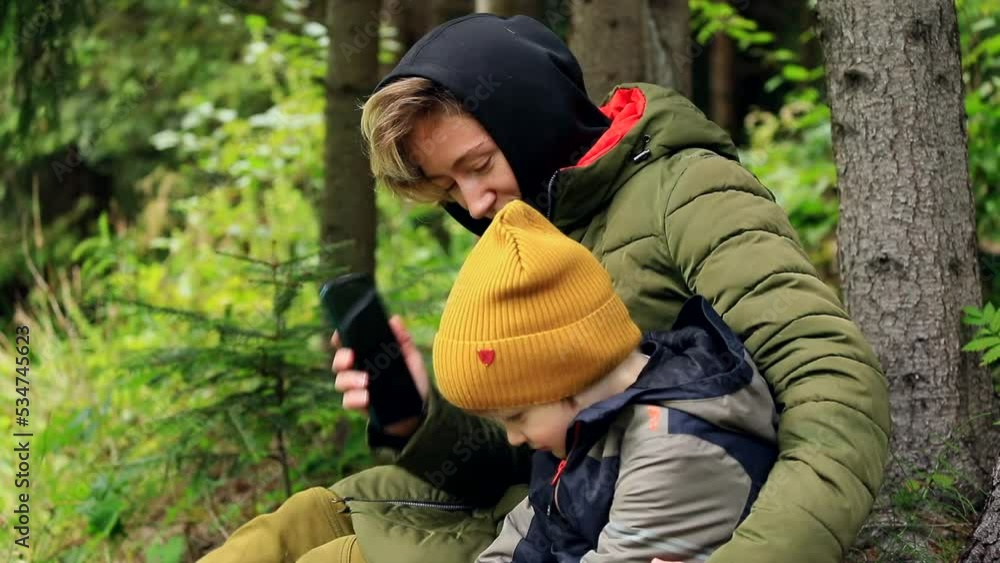 Young mother with happy little boy using smartphone for call resting in the mountains forest near the lake enjoying beautiful autumn nature. Internet connecting 5g. Traveling family lifestyle concept. Young mother with happy little boy using smartphone for call resting in the mountains forest near the lake enjoying beautiful autumn nature. Internet connecting 5g. Traveling family lifestyle concept.