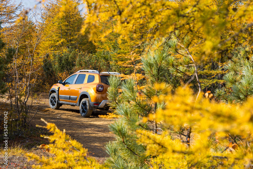 Car in the autumn forest. View through the branches of trees on an orange off-road car. Road trip in the wild. Beautiful northern nature. Fall season. Shallow depth of field and blurred foreground.