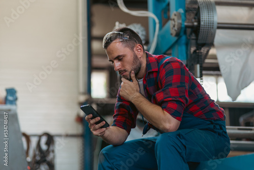 Young handsome bearded worker sitting on big machine and using smart phone. He is happy for success and cheering.