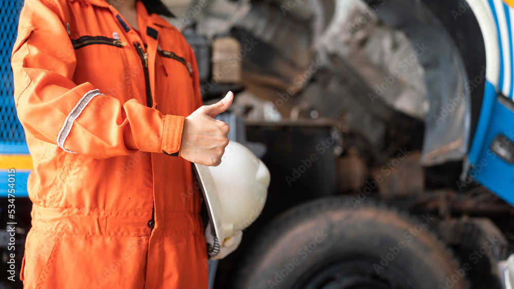 A repairman in orange coverall is thumbing up and holding white safety helmet with blurred background of garage workshop. Safe working practice in industrial concept.