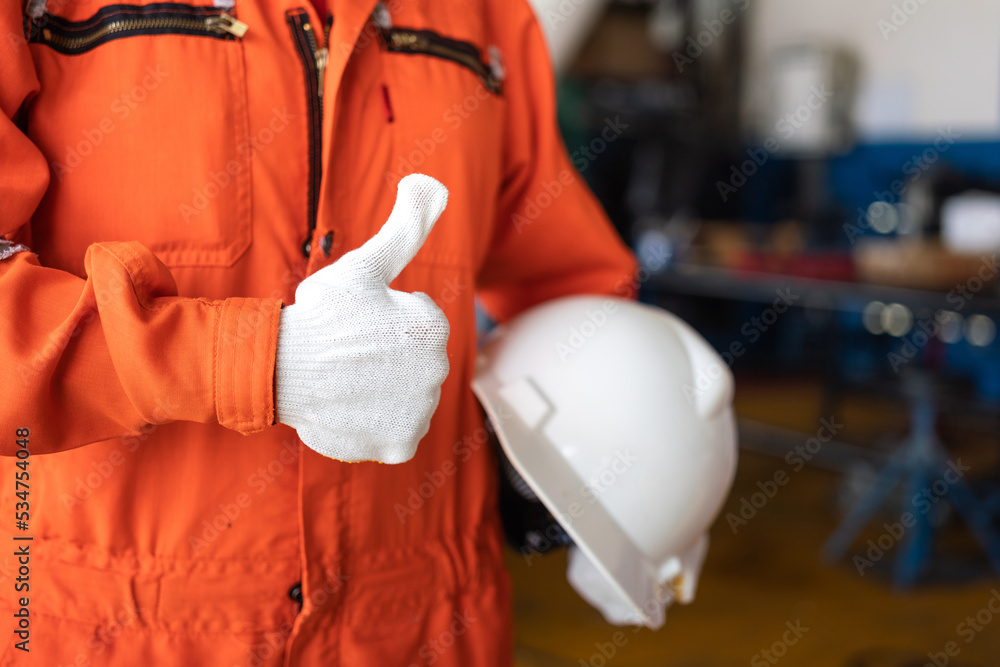 A repairman in orange coverall and white cotton glove is thumbing up and holding white safety helmet with blurred background of garage workshop. Safe working practice in industrial concept. A repairman in orange coverall and white cotton glove is thumbing up and holding white safety helmet with blurred background of garage workshop. Safe working practice in industrial concept.