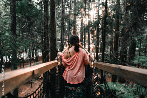 caucasian girl from the back in a red t-shirt and with a ponytail in her hair calmly walking along the wooden walkway between the trees in the forest, redwood treewalk, rotorua, new zealand