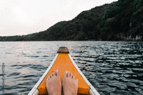 feet together resting on the prow of a white and orange wooden canoe heading into the forest navigating the lake waters at sunset, tarawera lake, new zealand photo