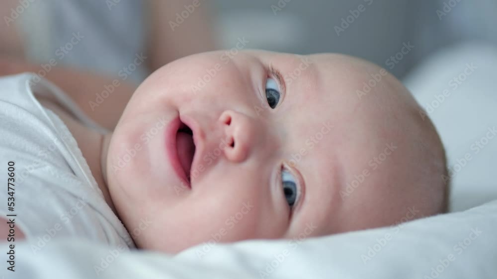 Baby girl with blue eyes looks around with smiling and happy expression lying on bed closeup. Excited baby enjoys parents care and laughs Baby girl with blue eyes looks around with smiling and happy expression lying on bed closeup. Excited baby enjoys parents care and laughs