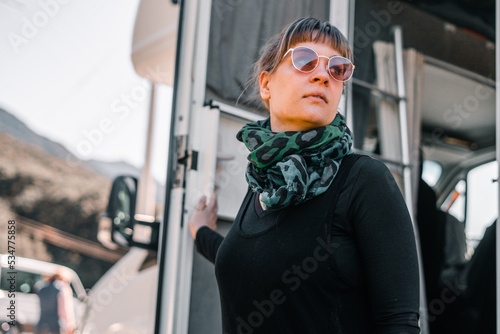 caucasian girl with bangs sunglasses and a large crumpled green scarf wrapped around her neck over a black t-shirt has just stepped out of a cozy motorhome at sunrise, matheson lake, new zealand