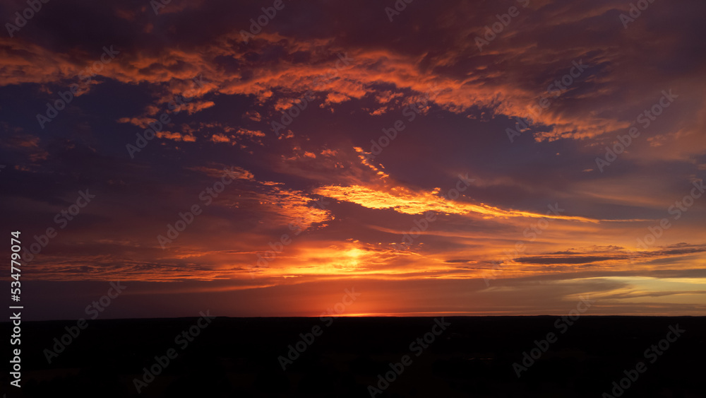 Clouds reflecting sunlight after sun has set below horizon, in hues of pink and orange over silhouetted, dark earth