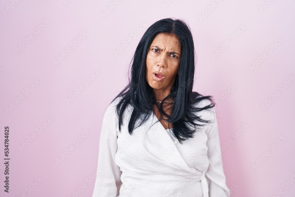 Mature hispanic woman standing over pink background in shock face, looking skeptical and sarcastic, surprised with open mouth