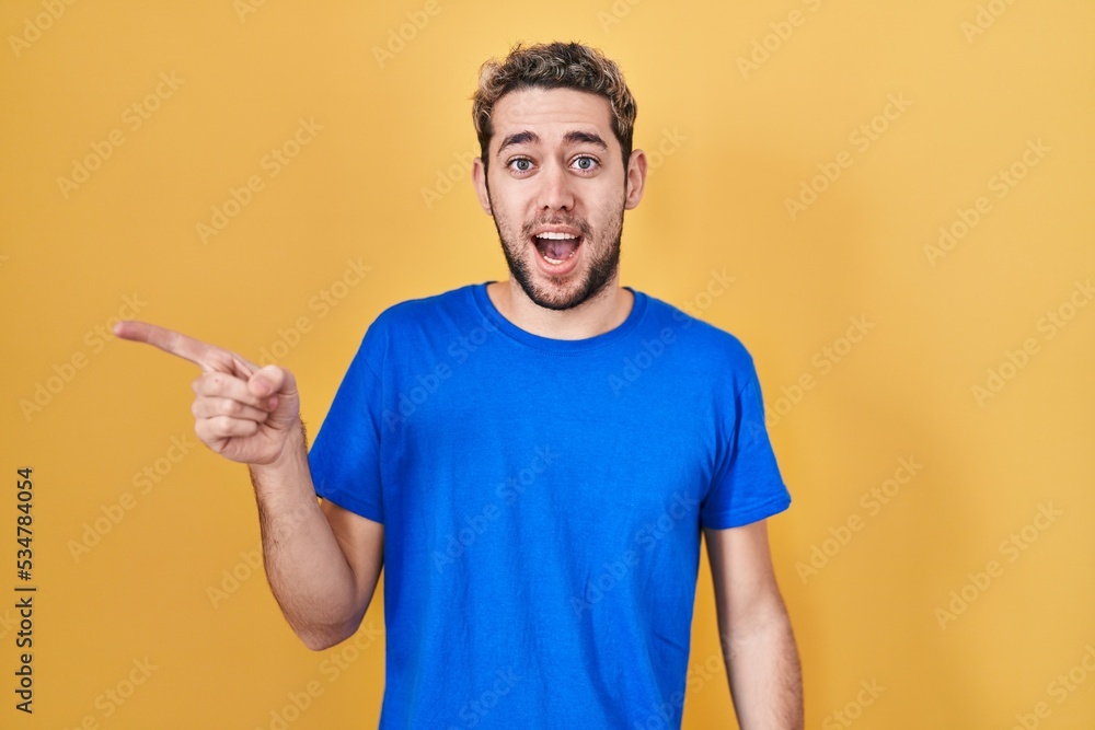 Hispanic man with beard standing over yellow background surprised pointing with finger to the side, open mouth amazed expression. Hispanic man with beard standing over yellow background surprised pointing with finger to the side, open mouth amazed expression.