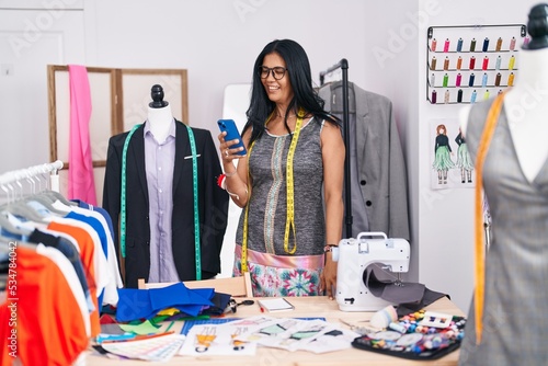 Middle age hispanic woman tailor smiling confident using smartphone at tailor shop