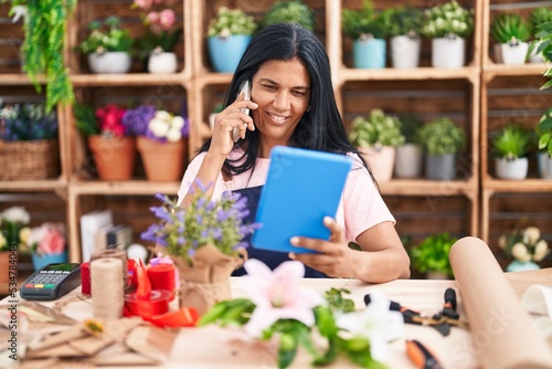 Middle age hispanic woman florist talking on smartphone using touchpad at florist