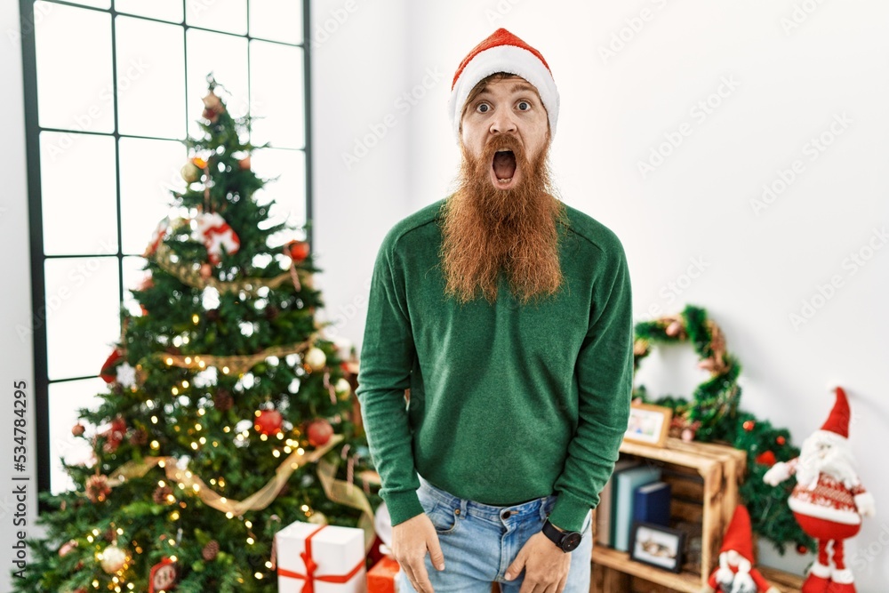 Redhead man with long beard wearing christmas hat by christmas tree afraid and shocked with surprise and amazed expression, fear and excited face. Redhead man with long beard wearing christmas hat by christmas tree afraid and shocked with surprise and amazed expression, fear and excited face.