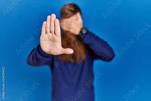 Redhead man with long beard wearing casual blue sweater over blue background covering eyes with hands and doing stop gesture with sad and fear expression. embarrassed and negative concept.